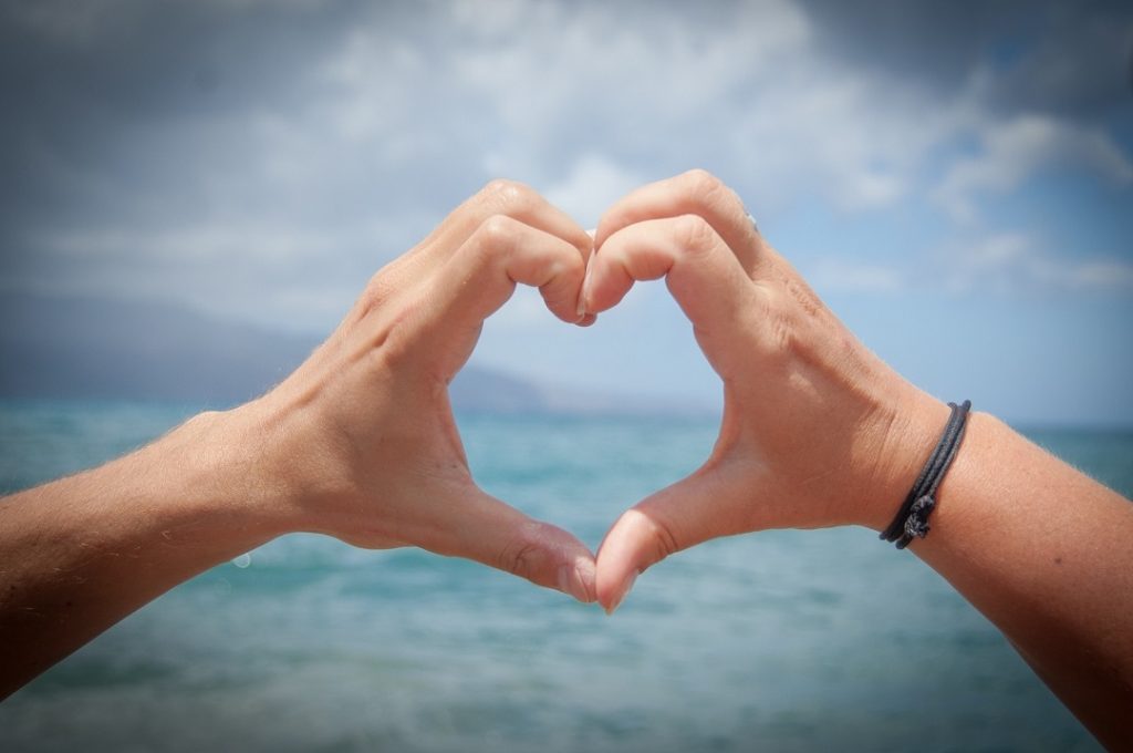 Two hands forming the shape of a heart in front of an ocean and sky background.